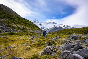 Two tourists are walking to the Kea Point Track in Mount Cook National Park, high rocky mountains and green grass in summer in New Zealand.