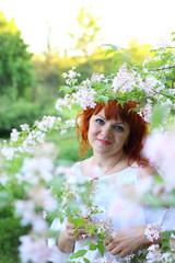 Naklejka premium beautiful red-haired Caucasian woman in a white dress enjoys flowering trees in the spring park 