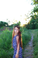 Fototapeta premium Cute Caucasian girl in a flower wreath walks at sunset in the park. The concept of summer and relaxation. Vacations and leisure for children. Nature and flowers.