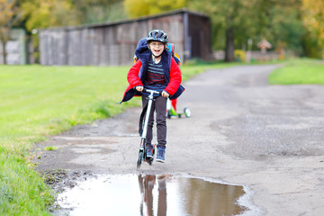cute little school kid boy riding on push scooter on the way to or from school. Schoolboy of 7 years driving through rain puddle. funny happy child in colorful fashion clothes and with helmet.