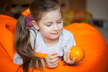 Fototapeta na wymiar Happy little girl wallows in an orange bean bag chair with a ripe orange fruit