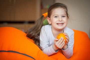 Fototapeta na wymiar Happy little girl wallows in an orange bean bag chair with a ripe orange fruit