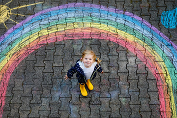 Happy little toddler girl in rubber boots with rainbow sun and clouds with rain painted with colorful chalks on ground or asphalt in summer. Cute child having fun. creative leisure