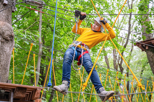 Boy in bright casual wear have a fun in activity with ladder rope park. Special equipment like protective helmet and gloves on the child. Outdoors, green trees around there.