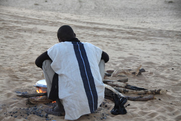 czarny mężczyzna w tradycyjnym stroju siedzący przy ognisku na pustyni - obrazy, fototapety, plakaty