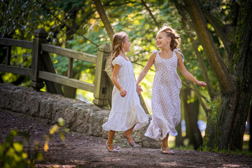 Two curly blonde girls in dresses run along the path of the summer park