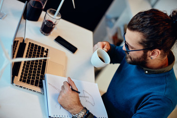 Young innovative dedicated bearded employee sitting in his modern office and drawing sketches of windmills and drinking coffee. Sustainable development concept.