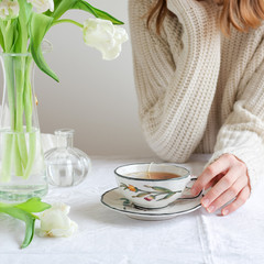 Detail of a young woman sitting at the table with white tulips in vase and drinking tea 