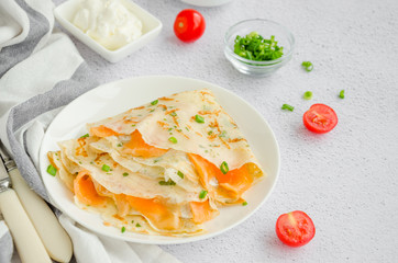 Traditional Russian thin pancakes or crepes with cream cheese, smoked salmon and green onions on a white plate on a light background. Holiday maslenitsa. Horizontal orientation.