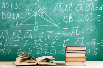 Education and reading concept - group of books on the wooden table, green blackboard with formulas on the background. Teachers desk