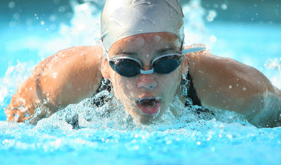 nadadora competindo de touca e óculos na piscina, nado borboleta na direção da câmera, ombros a...