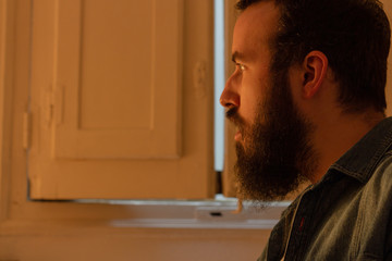 Man with a beard and denim shirt is sitting in room posing with surprised gesture.