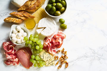 Cheese plate with brie, parmesan, cheddar and meat. Flatlay with variety of gourmet snacks, fruits and baguette on marble board. Copy space