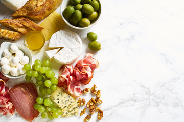 Cheese plate with brie, parmesan, cheddar and meat. Flatlay with variety of gourmet snacks, fruits and baguette on marble board. Copy space