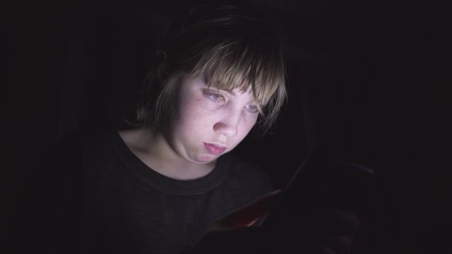 Sad/depressed teenager sitting on her bed in the dark while scrolling through her phone. The light from the phone is illuminating her face.