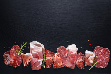 top view of set antipasti with sliced salami, prosciutto, ham, rosemary and dry peppers on black slate background, copy space