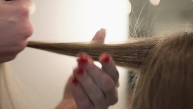 Hairdresser makes glowing curls with professional styling flat iron. Hairstylist works with young woman's long healthy hair. Creating of a wavy hairstyle. Preparing a client for an important event.