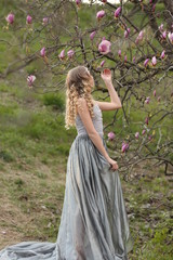 Obraz premium young blonde in a gray dress with a long train in the garden where pink magnolia blooms in spring