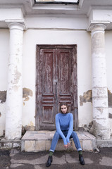Beautiful young girl is sitting on the stair in the blue jeans and blue sweater near old brown door. Street photo session. Street fashion.