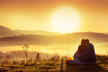 A couple of girl and guy are sitting in the early morning hugging and wrapping themselves in a plaid on a cliff overlooking the mountains and fog.