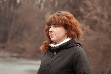 Young pretty woman with curly red hair in black jacket with a hood with a pensive look on the bridge in cloudy spring weather