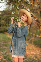  beautiful blonde girl with a bouquet of flowering branches in a straw hat and in a blue jeans jacket in a spring garden where trees bloom