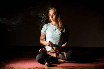 Fototapeta na wymiar Woman practicing yoga in a studio room sitting in lotus pose with the palo santo and resting bell