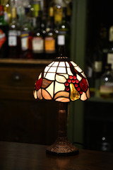Fototapeta na wymiar Retro table lamp with stained glass lampshade closeup on blurry background of bar. Glowing mosaic lamp in shape of dome. Vintage table-lamp with ornate lamp shade made of colorful glass pieces.