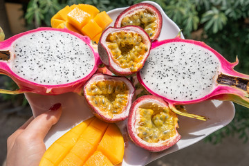 A mix of tropical fruits in the hands of a background of plants. Passion fruit, mango and dragon fruit in a plate top view.