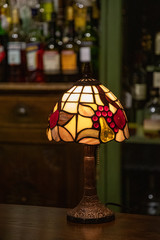 Fototapeta na wymiar Umbrella and lamp. Retro table lamp with stained glass lampshade closeup on blurry background of bar. Glowing mosaic lamp in shape of dome. Vintage table-lamp with ornate lampshade.