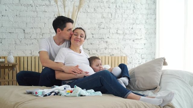 Young Father Touching Pregnant Belly of his Beautiful Wife while Sitting on the Bed at Home. Their Cute Little Son Playing Nearby. Pregnancy, Maternity, Preparation, Expectation and People Concept