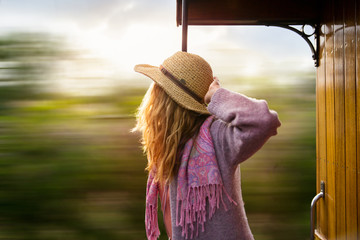 Young woman in straw hat traveling by retro wooden train. Sunset landscape, wind in blond hair. Girl smiling happily. Majorca, Spain. 