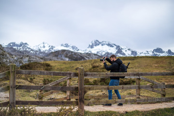 young man taking a picture from a bridge with scenic views of snowed mountains and green hills...