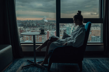Silhouette of a young girl in a white bathrobe which drinking her morning tea and looking throw the...