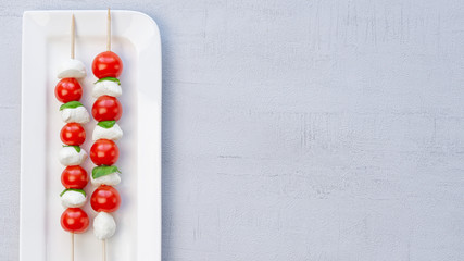 Naklejka na ściany i meble Caprese salad on a wooden stick in a white plate. Small mozzarella balls and cherry tomatoes. 