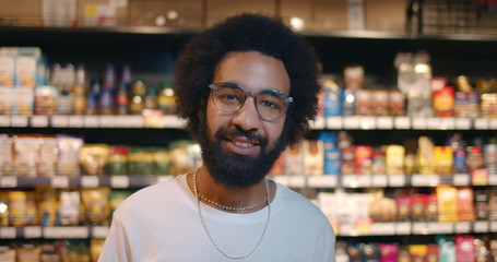 Close up of cheerful bearded guy in 30s smiling and looking to camera. Portrait of mature man in glasses wearing casual clothes and standing in supermarket. Concept of shopping.