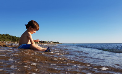 Four year old boy sitting by the sea at sunset, playing with water