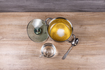 A metal pan with a glass lid contains millet grits, salt, sugar and a mug of water for cooking millet porridge on a wooden table