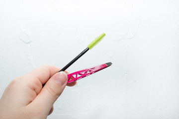 Eyelash brush, pink tweezers in a female hand on a uniform background. The concept of beauty and care for eyelashes and eyebrows.