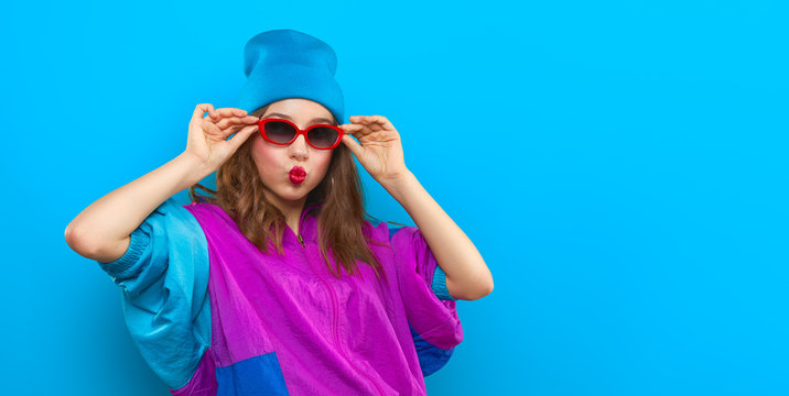 Cool teenager. Fashionable DJ girl in colorful trendy jacket and vintage retro sunglasses enjoys style of 80s � 90s vibes. Teenager Girl at disco party. Young fashion model on blue color background.