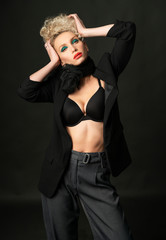 A beautiful blonde girl with an elegant hairstyle and large breasts, wearing a bra, trousers, scarf and a blazer, fashionable poses in the dark. Trendy, commercial and advertising design.