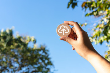 Diverse Asian female hand holding up wood disc with cloud computing upload icon - Cloud computer file transfer symbol with blue sky - Remote connectivity, data storage and digital security concepts