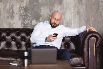Fototapeta premium Confident man corresponds with colleagues in the messenger on the phone. Stylish businessman conducts business online using a laptop. Business portrait