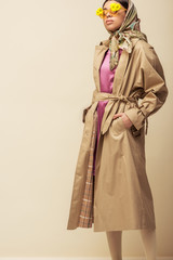 Fototapeta premium stylish african american girl in sunglasses with flowers, head scarf and trench coat standing with hands in pockets on beige