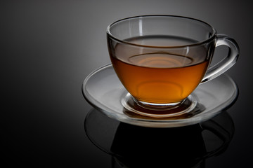 Glass cup of tea on a glass saucer with reflection, with a gradient of black and gray background. Concept, healthy and stylish lifestyle.