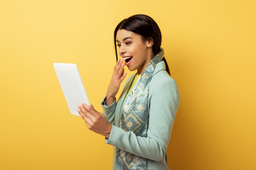 happy african american woman covering mouth and looking at digital tablet on yellow