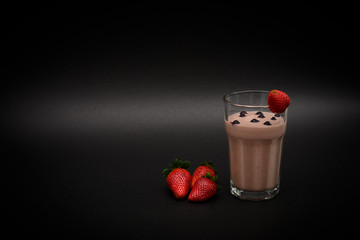 Juicy vitamin cold smoothie in a glass with one strawberry and blueberries on top and three strawberries next to a glass below. Dark background. Horizontal view. Copy space. Side view.