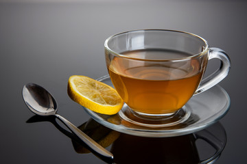 Glass cup of tea on a glass saucer with lemon and a spoon with reflection, with a gradient of black and gray background. Concept, healthy and stylish lifestyle.