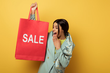 excited african american girl holding shopping bag with sale lettering on yellow