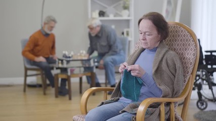 Old Caucasian woman knitting in armchair as blurred men playing chess at the background. Elderly retirees living in nursing home.